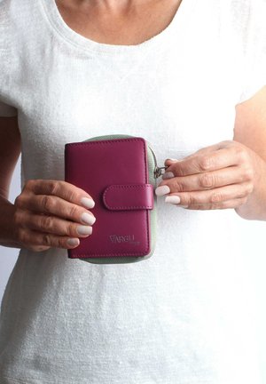 Kompakte Geldbörse aus burgunderfarbenem, veganem Leder mit grünem Akzent. Verfügt über einen Druckknopfverschluss und ein geprägtes Logo, gehalten in Händen vor einem weißen Hintergrund.