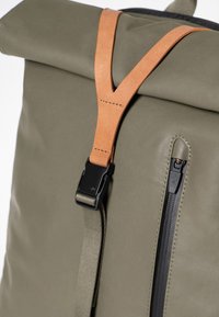 Zaino verde con chiusura a rotolo, dotato di una cintura in pelle beige e accessori neri, con una tasca laterale con zip e tessuto testurizzato.