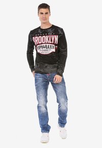 Schwarzes Langarm-T-Shirt mit "BROOKLYN"-Grafik in Rot und Weiß; kombiniert mit hellblauen, verwaschenen Jeans und weißen Sneakers.