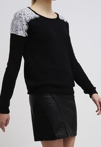 Femme portant un pull noir avec un détail en dentelle blanche sur l'épaule et une mini-jupe en cuir noir, sur un fond uni.