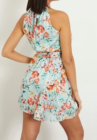 Žena v bezrukávových světle modrých květovaných šatech s volány na spodním lemu a páskem v pase, zobrazená zezadu na jednobarevném pozadí.