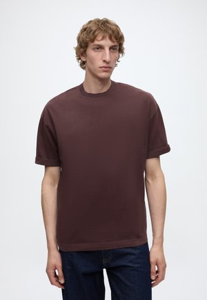 Junger Mann mit lockigen hellbraunen Haaren, der ein schlichtes dunkelbraunes Kurzarmshirt und dunkelblaue Jeans trägt, steht vor einem schlichten weißen Hintergrund.