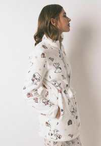 Biała polarowa szlafrok z wielokolorowymi wzorami z Psotnika i motywami świątecznymi, zapinany na zamek, z wysokim kołnierzem, paskiem w talii i bocznymi kieszeniami.