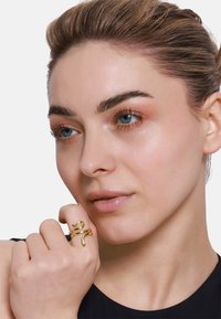 Anello d'oro a forma di serpente con un design testurizzato, indossato su una mano. Primo piano di un volto con pelle liscia e trucco neutro su uno sfondo bianco.