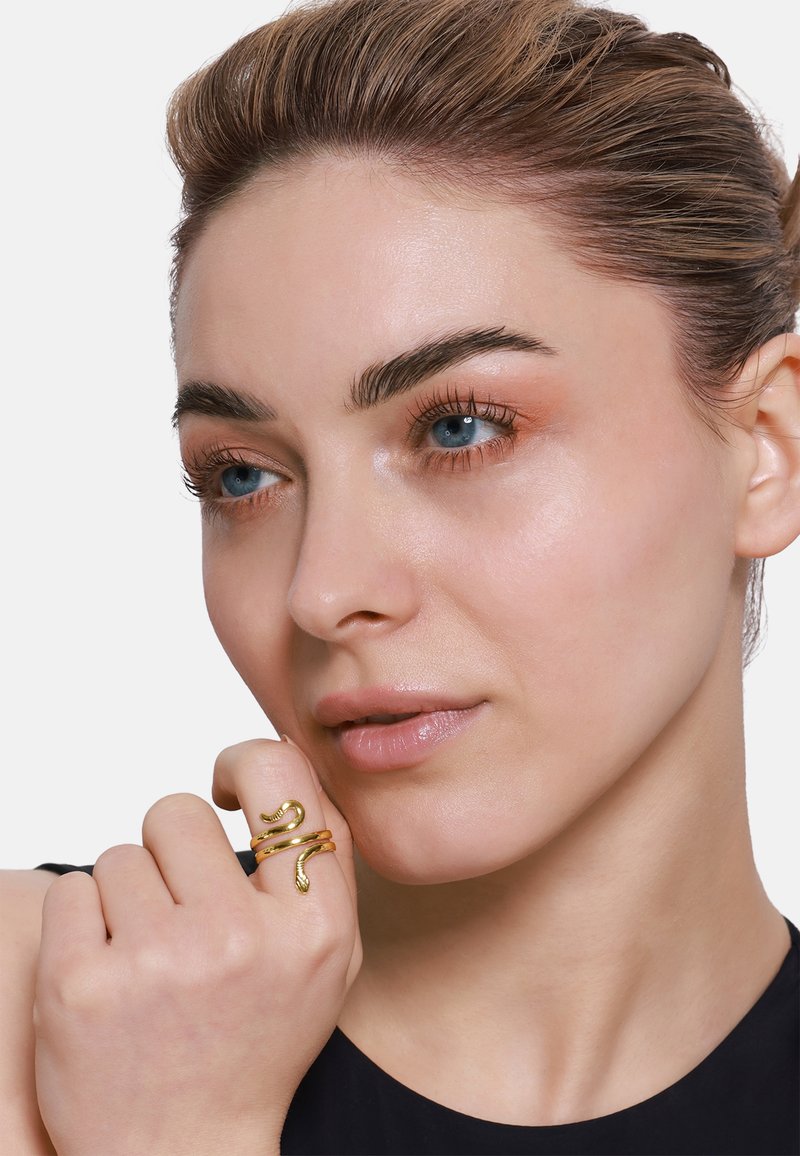 Anello d'oro a forma di serpente con un design testurizzato, indossato su una mano. Primo piano di un volto con pelle liscia e trucco neutro su uno sfondo bianco.