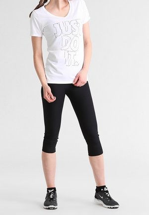 Vrouw draagt een witte "JUST DO IT." t-shirt, zwarte cropped leggings en zwarte hardloopschoenen, staand tegen een effen achtergrond.