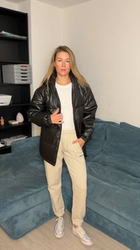 Veste matelassée noire avec poignets élastiques, portée sur une chemise blanche et un jogging beige. Le tissu a une finition brillante et une fermeture éclair à l'avant.