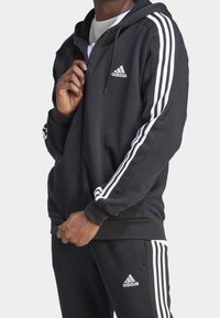 Felpa nera con zip realizzata in tessuto morbido, con dettagli bianchi a tre strisce sulle maniche e un piccolo logo sul petto. Design con cappuccio.