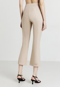 Pantaloni beige a palazzo con vita alta, texture liscia e silhouette aderente. Abbinati a tacchi neri con cinturini.