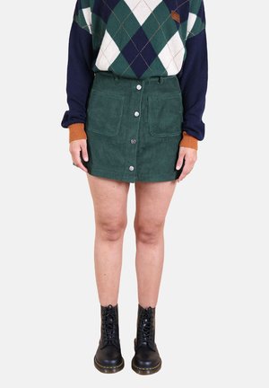 Mini-jupe en velours côtelé vert avec des boutons sur le devant et des poches, assortie à un pull à motifs dans des tons bleu marine et vert, ainsi qu'à des bottines noires à lacets.