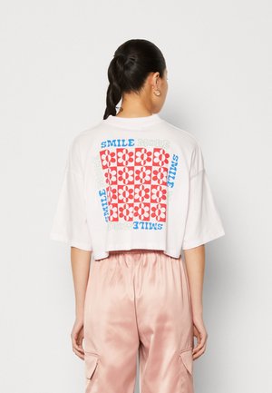 Valkoinen lyhythihainen cropped t-paita, jossa on värikäs kukkakuosi ja selässä teksti "Hymyile enemmän" sinisellä ja vihreällä.