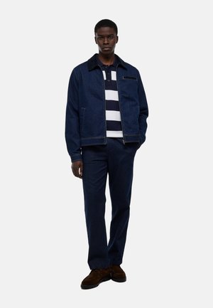 Jeune homme debout portant une veste en denim bleu foncé, un pantalon assorti, un polo rayé bleu marine et blanc, et des chaussures marron sur fond blanc.