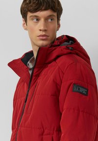 Czerwona pikowana kurtka z wysokim kołnierzem i kapturem. Z czarnymi akcentami, kieszenią na piersi na zamek oraz przeszyciami tworzącymi fakturę. Logo na rękawie.