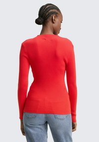 Roter gerippter Strickpullover mit langen Ärmeln, taillierter Passform und rundem Ausschnitt, kombiniert mit hellblauen Jeans.
