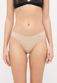 Béžové tangá s čipkovým lemovaním, vyrobené z mäkkého materiálu a s elastickým pásom, na ktorom je logo Tommy Hilfiger.