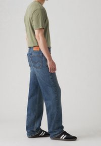 Man som bär en olivgrön t-shirt, lösa blå jeans och svarta sneakers med vita ränder, stående på sidan mot en neutral bakgrund.