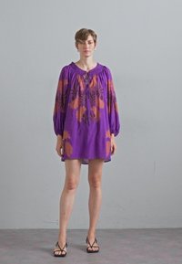 Paarse jurk met lange mouwen, versierd met oranje bloemenpatronen en een ronde hals met een striksluiting. Draagt een paar zwarte sandalen.