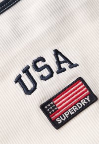 Weißer Stoff mit gerippter Textur zeigt blau gestickten "USA"-Text und ein rechteckiges Patch mit einer rot-blauen amerikanischen Flagge und "SUPERDRY."