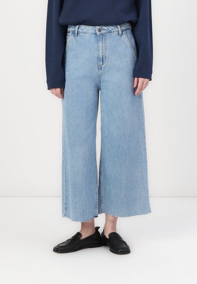 Jean en denim large, de couleur bleu clair, à taille haute, avec un ourlet brut et une légère effilochure aux poignets. Porté avec des mocassins noirs.