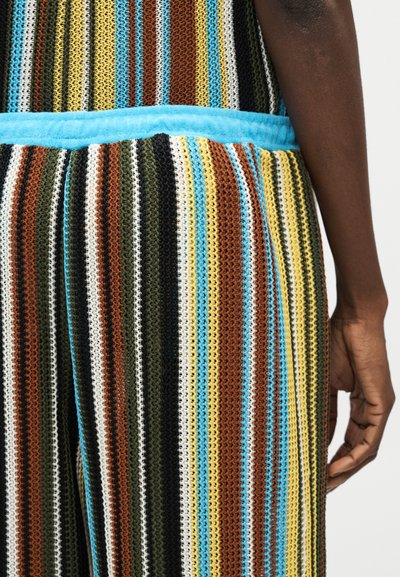 Κοντινή λήψη του χεριού ενός ατόμου δίπλα σε πλεκτό ρούχο με κάθετες ρίγες σε μπλε, κίτρινο, καφέ, λευκό, μαύρο και πράσινο, με ένα ανοιχτό μπλε ζωνάρι στη μέση.