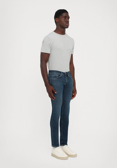 Uomo in piedi di fronte, leggermente rivolto a destra, con indosso una maglietta a maniche corte grigia chiara, jeans slim blu scuro e sneakers bianche su uno sfondo neutro.