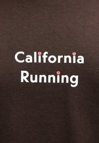Λευκό κείμενο "California Running" σε σκούρο καφέ ύφασμα με μικρά ροζ σχήματα λουλουδιών που αντικαθιστούν τις τελείες πάνω από τα γράμματα i.