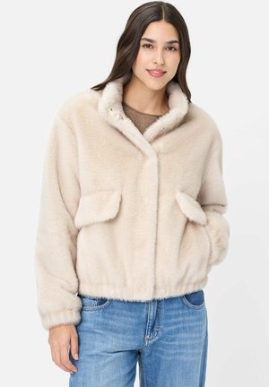 Young woman wearing a beige faux fur jacket with flap pockets and blue jeans against a plain white background.