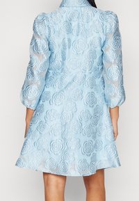 Kvinna iklädd en ljusblå klänning med ett blommigt spetsmönster och trekvarts genomskinliga ärmar, visad bakifrån.