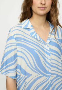 Blusa a maniche corte in tessuto morbido, caratterizzata da un motivo a onde blu e bianche, con colletto a punta e chiusura frontale con bottoni.