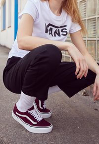 Sneakers in suede color granata con accenti bianchi, indossate con calzini bianchi, pantaloni neri corti e una t-shirt bianca con un logo nero.