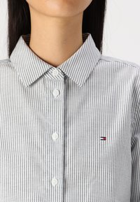 Camicia button-up in cotone a righe grigio e bianco, con colletto a punta, bottoni bianchi e un piccolo logo marchiato rosso, bianco e blu.