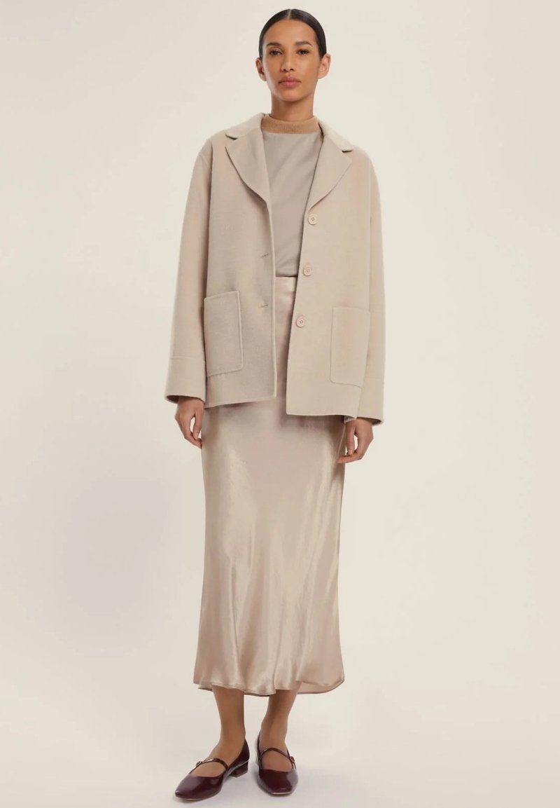 Blazer in lana beige con ampie tasche, indossato sopra un vestito in satin crema. Ballerine marroni completano l'outfit. Texture morbida e linee pulite.