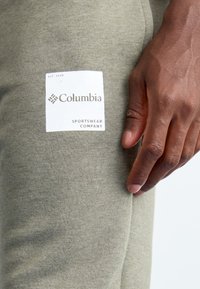 Prim-plan al unei mâini odihnindu-se pe un material verde măsliniu deschis cu un patch al logo-ului Columbia Sportswear Company alb.