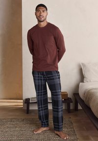 Langärmliges burgunderfarbenes Oberteil mit einer Brusttasche, kombiniert mit schwarz-grauen karierten Hosen. Glatter Stoff, lockere Passform, barfuß auf einem strukturierten Teppich.