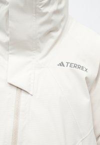 Artimas baltos Terrex prekės ženklo striukės kadras, su priekiniais užtrauktuku ir uždengta klape.