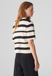 Pull court à manches courtes en crème texturé avec des rayures noires. Il présente un col rond et une coupe décontractée. Un pantalon foncé est visible en dessous.
