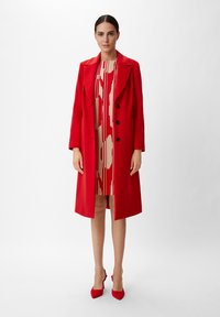 Roter langer Mantel mit schwarzen Knöpfen, über einem beigen Kleid mit roten Mustern getragen, kombiniert mit roten spitz zulaufenden Schuhen. Glattes Material, strukturiertes Design.