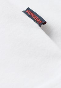Tissu blanc avec une petite étiquette noire et rouge cousue portant la mention « SUPERDRY » attachée au bord.