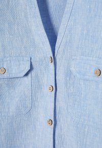 Chemise en lin bleu clair avec deux poches à rabat sur le devant et des boutons beiges au centre.