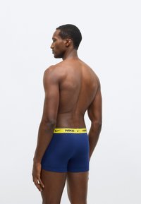 Tmavomodré dielové boxerky s žltým pásom značky Nike, ktoré majú priliehavý strih a jemnú textúru. Modelka je zobrazená zozadu, kde je vidieť dizajn.