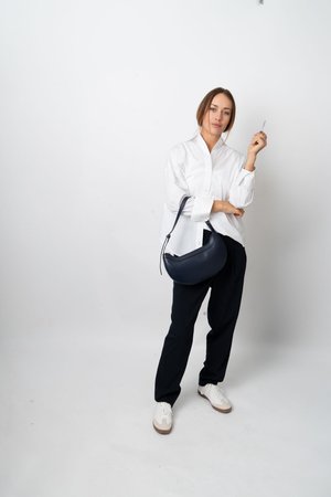 Femme en chemise blanche, pantalon noir et baskets blanches tient un sac à main bleu marine et un petit objet devant un fond clair uni.