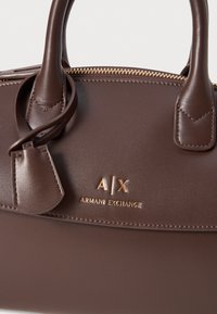 Sac à main en simili cuir marron avec doubles poignées, fermeture éclair dorée, logo embossé Armani Exchange et un petit détail de tag de bagage.