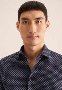 Marineblauwe blouse met een geometrisch patroon in wit en lichtblauw, voorzien van een knoopsluiting en een gladde textuur.