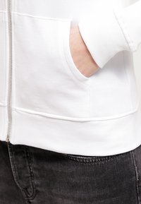 Sudadera blanca con cremallera, con puños y dobladillo acanalados, que presenta un bolsillo lateral. La tela parece suave y ligera, combinada con jeans oscuros.