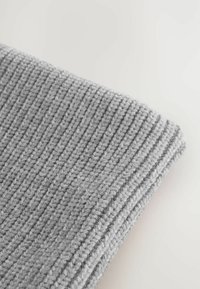 Grau gestrickter Stoff mit einem strukturierten, gerippten Muster. Das Material wirkt weich und dick, am einen Rand umgeschlagen.