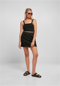 Zwarte crop top met dikke banden, zwarte elastische rok en zwarte sandalen met een brede band. Model poseert tegen een effen grijze achtergrond.