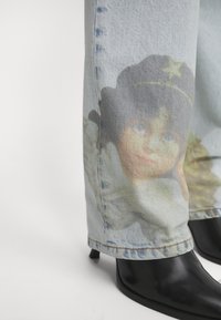 Jeans in denim blu chiaro con un ritratto stampato di un volto vicino alla caviglia, abbinati a stivali neri con tacco a punta.
