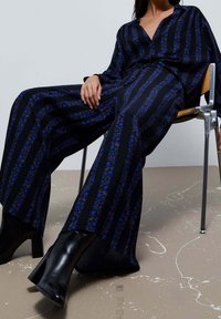 Schwarzer und blauer gestreifter Jumpsuit mit floralem Muster, langen Ärmeln, tiefem V-Ausschnitt, weitem Bein, kombiniert mit schwarzen Knöchelboots, sitzt auf einem Stuhl.