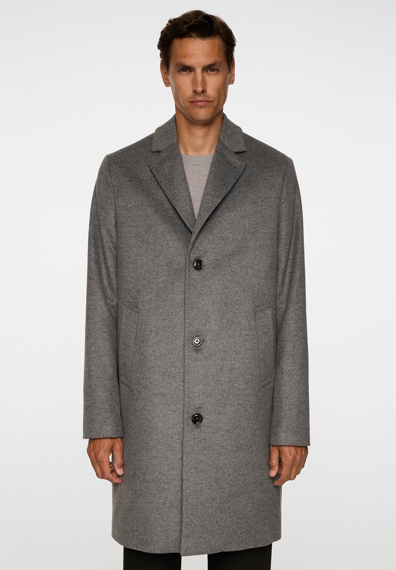 Uomo che indossa un lungo cappotto grigio di lana con colletto a risvolto e tre bottoni neri sopra un maglione grigio chiaro, in piedi su uno sfondo bianco.
