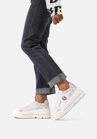 Sneakers bianchi con un accento beige testurizzato, dotati di dettagli metallici, logo rotondo e suola spessa. Indossati con jeans scuri arrotolati.